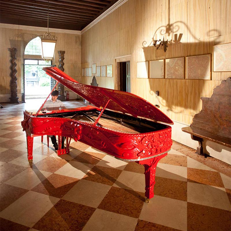 Red Steinway grand piano intricately carved with Māori designs, being played by a young man inside the Palazzo Loredan dell’Ambasciatore in Venice.  