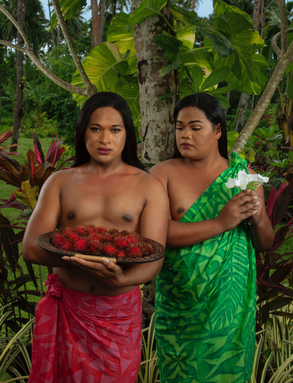 Two Fa’afafine (Samoa’s third gender) stand in front of foliage, one holds bowl of rambutans and the other a white flower.