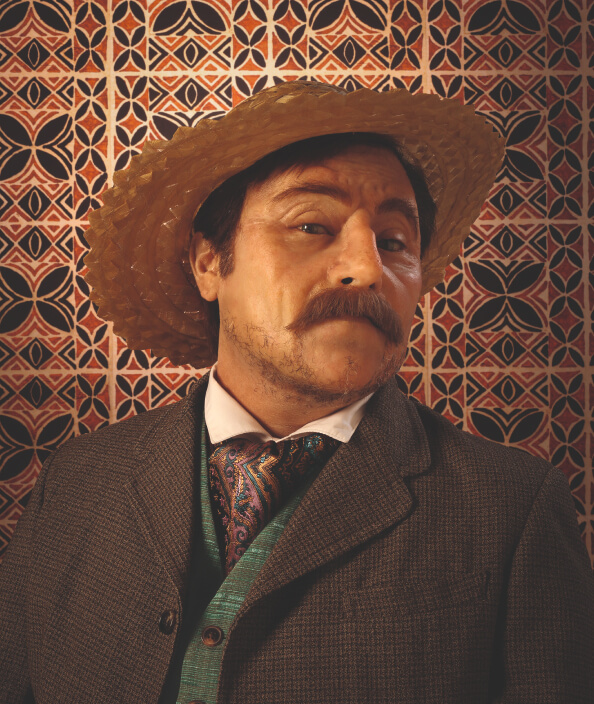Head shot of Yuki Kihara wearing a three-piece suit, silk cravat and Sāmoan straw hat in front of a siapo backdrop.