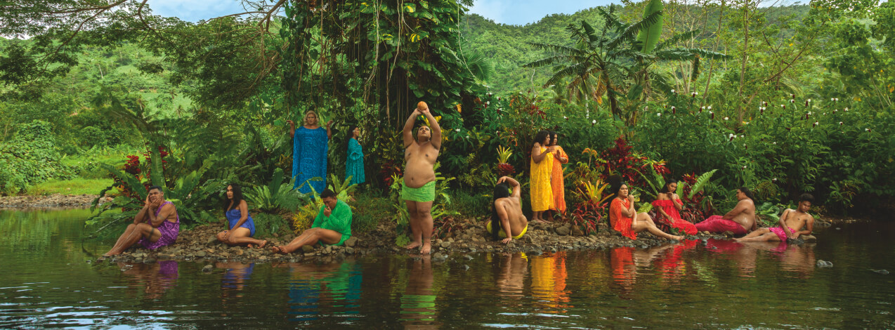 Thirteen Fa‘afafine wearing rainbow-coloured Pacific dresses on a rocky riverbed in front of dense tropical foliage