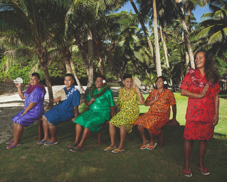 Six Fa’atama in front of palm trees wear a different colour of the rainbow and hold a conch shell and woven fans.