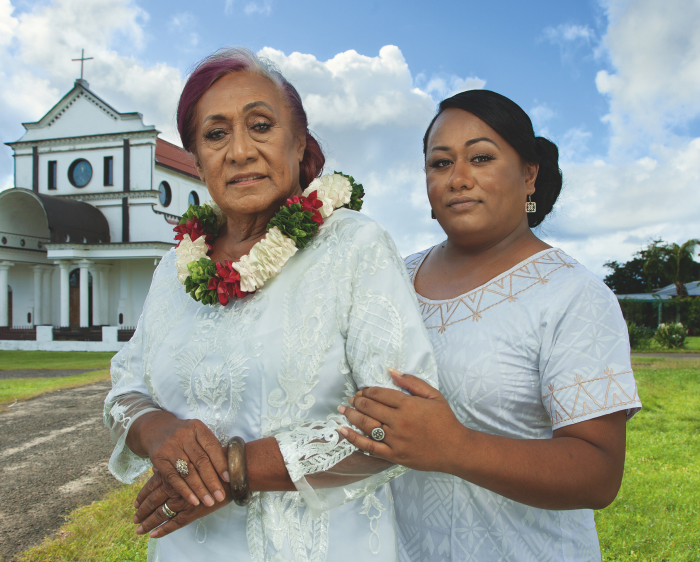 Two Fa‘afafine (Sāmoa’s third gender) in traditional white church attire in front of a church. One wears a colourful flower necklace.