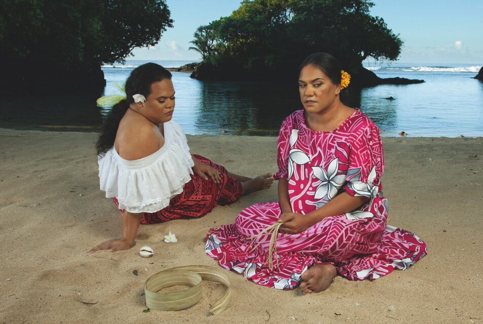 Two Fa‘afafine (Sāmoa’s third gender) on a beach. One holds dried pandanus strips and the other has a flower in their hair.