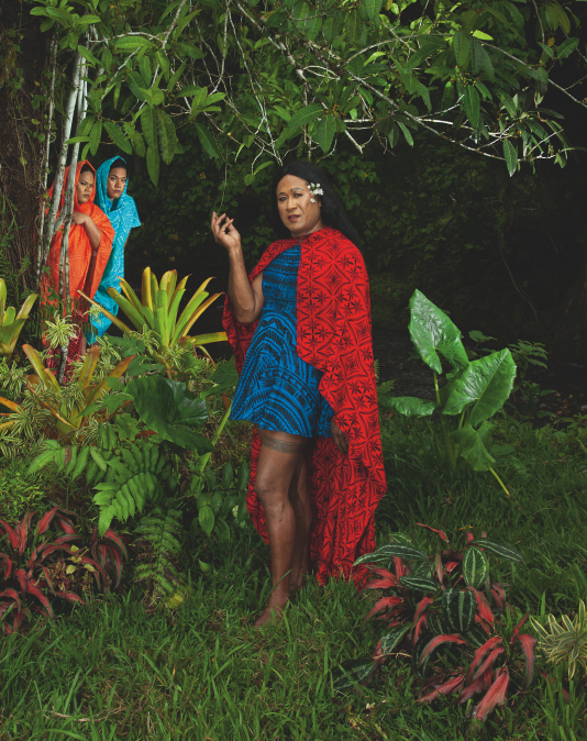 A Fa‘afafine (Samoa’s third gender) stands amongst tropical foliage, watched by two figures behind a tree.